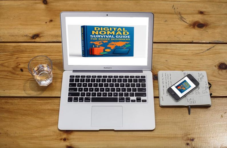 Op de afbeelding: Een laptop toont de boekomslag "DIGITAL NOMAD SURVIVAL GUIDE FOR BROKE BEGINNERS". Een smartphone en notitieboek liggen ernaast. Een glas water staat links op een houten oppervlak. De sc&egrave;ne suggereert een werkplek.