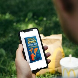 Op de afbeelding: Een smartphone toont de omslag van een boek met de titel "DIGITAL NOMAD SURVIVAL GUIDE FOR BROKE BEGINNERS". De omslag toont een wereldkaart, een koffer en een laptop. De telefoon wordt vastgehouden tegen een groene gras achtergrond, met een fastfoodmaaltijd op de achtergrond.