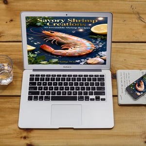 May include: A laptop displays a cookbook cover with a large shrimp and the title "Savory Shrimp Creations." A smartphone and notebook are nearby. A glass of water sits to the left on a wooden surface.