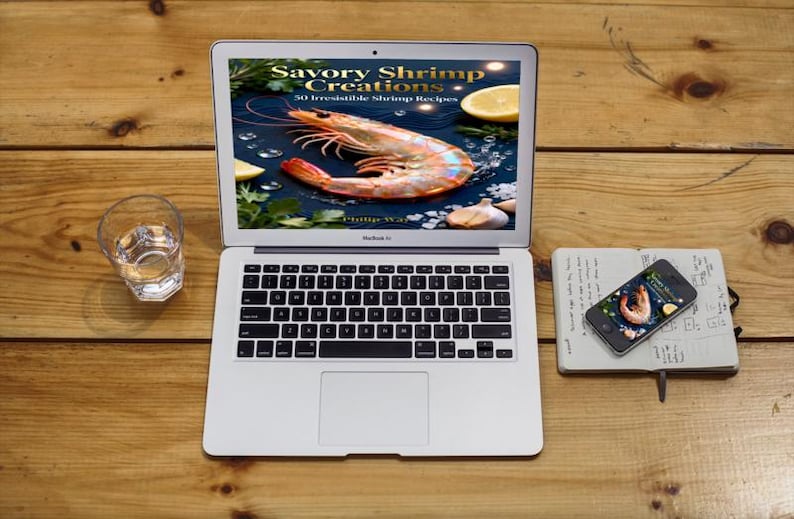 May include: A laptop displays a cookbook cover with a large shrimp and the title "Savory Shrimp Creations." A smartphone and notebook are nearby. A glass of water sits to the left on a wooden surface.