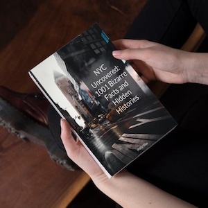 May include: A book titled "NYC Uncovered: 1001 Bizarre Facts and Hidden Histories" is held open. The cover features a grayscale image of a city street with tall buildings. The book is held by a person wearing black clothing.