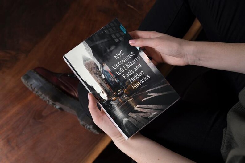 May include: A book titled "NYC Uncovered: 1001 Bizarre Facts and Hidden Histories" is held open. The cover features a grayscale image of a city street with tall buildings. The book is held by a person wearing black clothing.