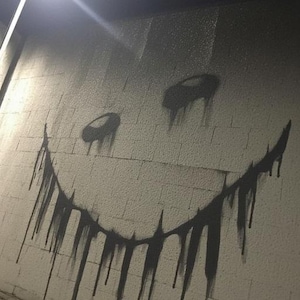 May include: A black and gray graffiti smiley face with dripping paint on a white brick wall. The artwork is illuminated by a bright overhead light, creating shadows and a stark contrast.