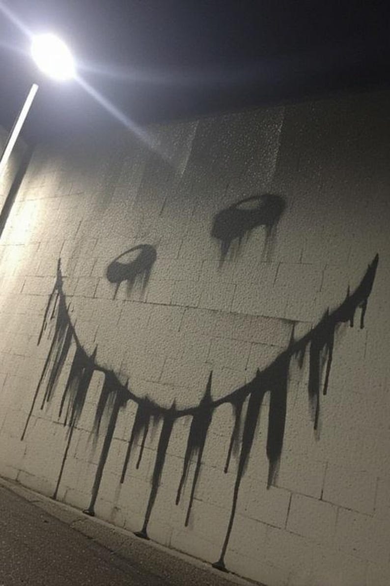 May include: A black and gray graffiti smiley face with dripping paint on a white brick wall. The artwork is illuminated by a bright overhead light, creating shadows and a stark contrast.