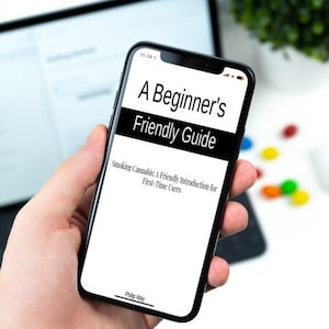 Op de afbeelding: Een zwarte smartphone toont de tekst "A Beginner's Friendly Guide" met de ondertitel "Smoking Cannabis: A Friendly Introduction for First-Time Users." De telefoon wordt in een hand gehouden, met een vage achtergrond van een tablet en groen.