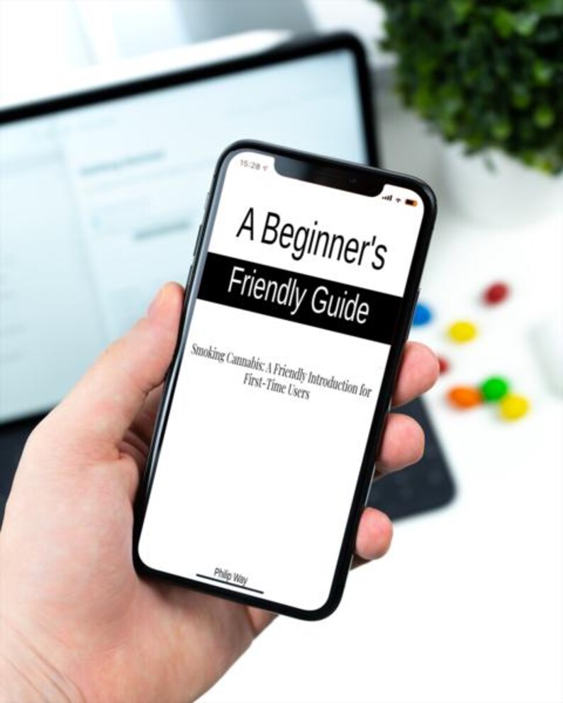 Op de afbeelding: Een zwarte smartphone toont de tekst "A Beginner's Friendly Guide" met de ondertitel "Smoking Cannabis: A Friendly Introduction for First-Time Users." De telefoon wordt in een hand gehouden, met een vage achtergrond van een tablet en groen.