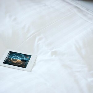 May include: A white tablet displaying the text "Code Breaker" and a futuristic graphic, resting on a white bedsheet. The tablet is positioned on the left side of the frame, with a pillow in the upper right corner.