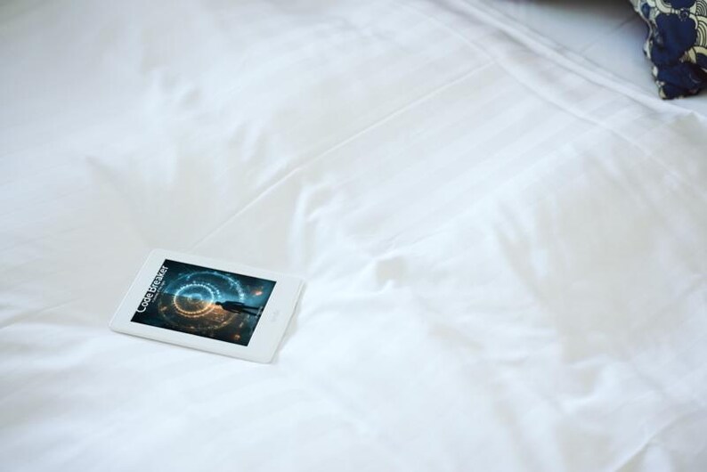 May include: A white tablet displaying the text "Code Breaker" and a futuristic graphic, resting on a white bedsheet. The tablet is positioned on the left side of the frame, with a pillow in the upper right corner.