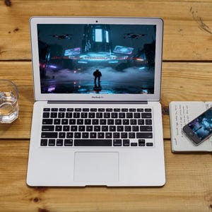 K&ouml;nnte beinhalten: Ein silberner Laptop mit schwarzer Tastatur zeigt eine futuristische Stadtlandschaft. Ein Glas Wasser und ein Smartphone auf einem Notizblock liegen neben dem Laptop auf einer Holzoberfl&auml;che. Der Smartphone-Bildschirm spiegelt die Anzeige des Laptops wider.