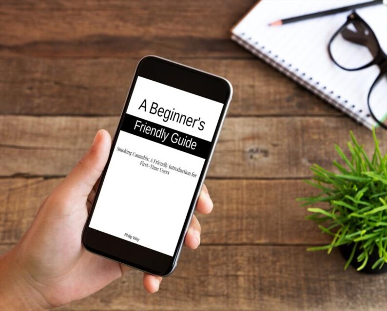 Op de afbeelding: Een smartphone toont de tekst "A Beginner's Friendly Guide". De ondertitel van de gids luidt "Smoking Cannabis: A Friendly Introduction for First-Time Users". De telefoon wordt in een hand gehouden, met een houten achtergrond.