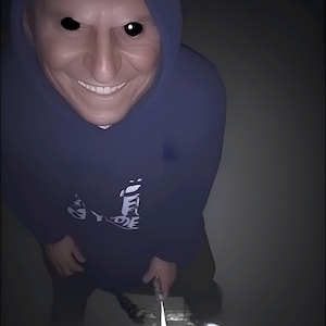 May include: A digitally altered image of a person wearing a dark blue hooded sweatshirt with a white graphic. The person has a distorted face with black eyes and a wide smile, holding a knife.