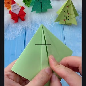 May include: A person is folding a green paper square into a pyramid shape. The paper is folded in half, and the person is pressing down on the fold to make a crease.