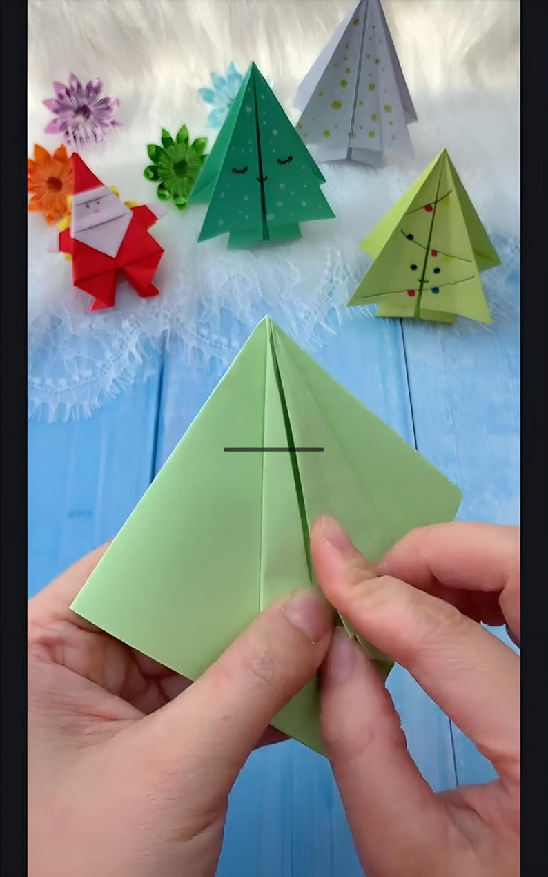 May include: A person is folding a green paper square into a pyramid shape. The paper is folded in half, and the person is pressing down on the fold to make a crease.