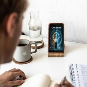May include: A smartphone displaying the "Code Breaker" app is propped on a wooden stand. A person is writing in a notebook. A gray mug with an apple logo sits on a coaster, next to a glass water bottle.