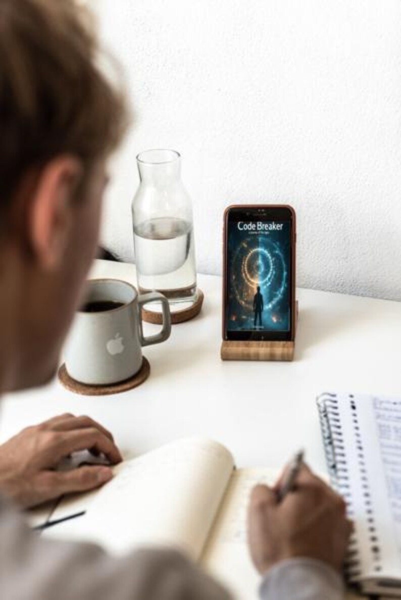 May include: A smartphone displaying the "Code Breaker" app is propped on a wooden stand. A person is writing in a notebook. A gray mug with an apple logo sits on a coaster, next to a glass water bottle.