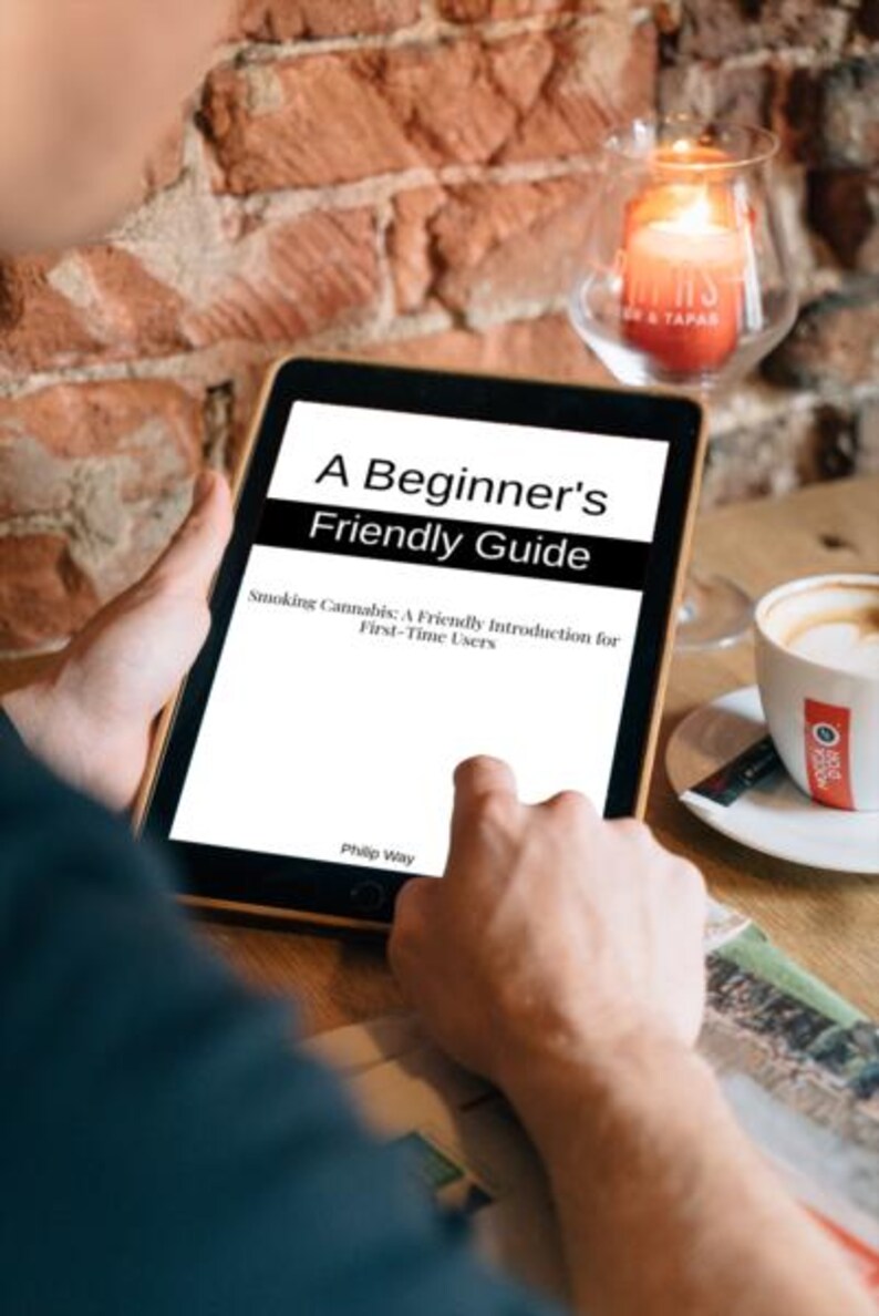 Op de afbeelding: Een tablet met de tekst "A Beginner's Friendly Guide" en de ondertitel "Smoking Cannabis: A Friendly Introduction for First-Time Users". Op de achtergrond een kopje koffie en een brandende kaars in een glazen houder, tegen een bakstenen muur.