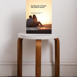 May include: A book titled "Decoding Men: A Straight Talk Guide for Women" is displayed on a small, white, round-top stool with wooden legs. The book cover features a silhouette of a couple at sunset. The stool sits on a wooden floor.
