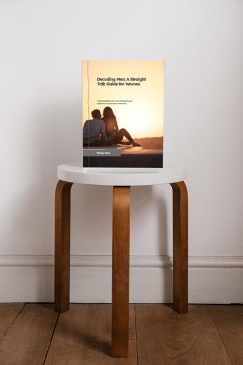 May include: A book titled "Decoding Men: A Straight Talk Guide for Women" is displayed on a small, white, round-top stool with wooden legs. The book cover features a silhouette of a couple at sunset. The stool sits on a wooden floor.
