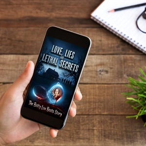 K&ouml;nnte beinhalten: Ein Smartphone zeigt das Cover des Buches "Love, Lies and Lethal Secrets". Das Cover zeigt ein Spukhaus, ein Medaillon mit dem Portr&auml;t einer Frau und den Text "The Betty Lou Beets Story". Das Telefon liegt auf einem Holzgrund.