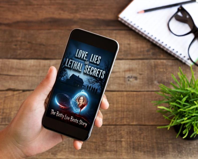K&ouml;nnte beinhalten: Ein Smartphone zeigt das Cover des Buches "Love, Lies and Lethal Secrets". Das Cover zeigt ein Spukhaus, ein Medaillon mit dem Portr&auml;t einer Frau und den Text "The Betty Lou Beets Story". Das Telefon liegt auf einem Holzgrund.
