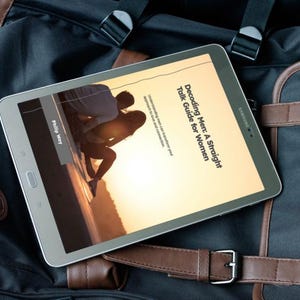 May include: A silver Samsung tablet displaying the cover of a book titled "Decoding Men: A Straight Talk Guide for Women." The tablet rests on a black and brown backpack with leather straps.