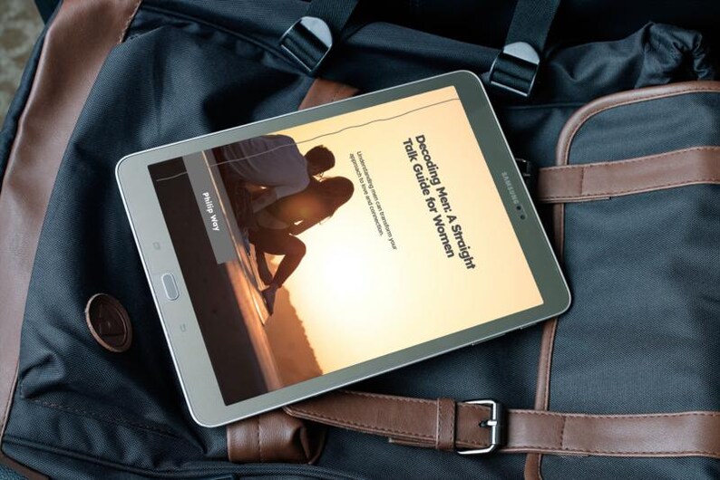 May include: A silver Samsung tablet displaying the cover of a book titled "Decoding Men: A Straight Talk Guide for Women." The tablet rests on a black and brown backpack with leather straps.