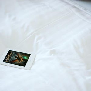 Op de afbeelding: Een witte e-reader met de cover van een boek getiteld "Eldoria" ligt op een wit laken. De boekomslag toont een fantasie-illustratie met een draak en een gloeiende bol. Een kussen met patroon is op de achtergrond.