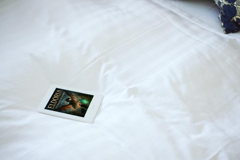 Op de afbeelding: Een witte e-reader met de cover van een boek getiteld "Eldoria" ligt op een wit laken. De boekomslag toont een fantasie-illustratie met een draak en een gloeiende bol. Een kussen met patroon is op de achtergrond.