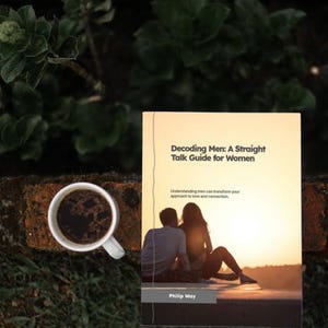 May include: A book titled "Decoding Men: A Straight Talk Guide for Women" with a cover image of a couple at sunset. A white coffee cup filled with dark liquid sits to the left of the book, with a background of greenery and brick.
