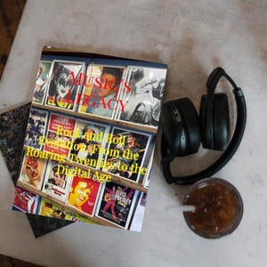 K&ouml;nnte beinhalten: Ein Buch mit dem Titel "Music's Legacy" mit Bildern von Rock'n'Roll-K&uuml;nstlern, neben schwarzen Kopfh&ouml;rern und einem Glas Eiskaffee mit Strohhalm. Der Untertitel des Buches lautet "Rock and Roll Evolution From the Roaring Twenties to the Digital Age."