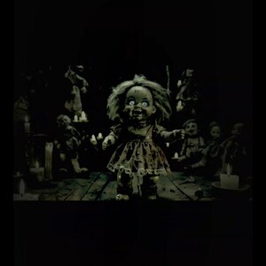 Puede incluir: Una mu&ntilde;eca espeluznante con ojos blancos y una sonrisa siniestra se encuentra en una habitaci&oacute;n con poca luz. La mu&ntilde;eca lleva un vestido hecho jirones y est&aacute; rodeada de otras mu&ntilde;ecas y velas, creando una atm&oacute;sfera espeluznante. La escena evoca un tema de terror.