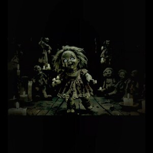 Puede incluir: Una mu&ntilde;eca espeluznante con ojos blancos y una sonrisa siniestra se encuentra en una habitaci&oacute;n con poca luz. La mu&ntilde;eca lleva un vestido hecho jirones y est&aacute; rodeada de velas y otras mu&ntilde;ecas. La escena evoca un tema de terror.