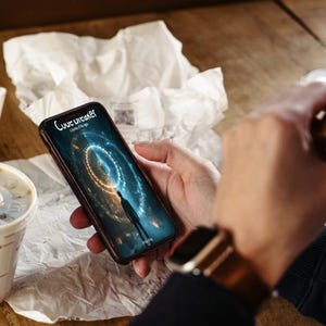 May include: A smartphone displaying a digital graphic with the text "Love Breaker: A game of the age." The phone is held in a person's hand, with a watch on their wrist. A McDonald's cup and food are also visible.