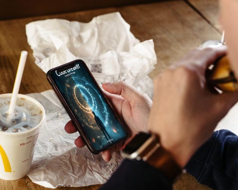 May include: A smartphone displaying a digital graphic with the text "Love Breaker: A game of the age." The phone is held in a person's hand, with a watch on their wrist. A McDonald's cup and food are also visible.