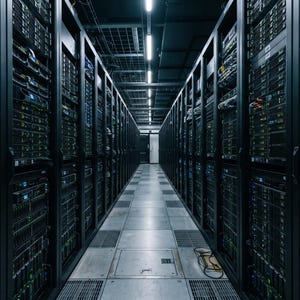 May include: A server room featuring two parallel rows of black server racks filled with stacked hardware units. Each unit displays small, glowing green and blue status indicator lights, creating a symmetrical corridor of technology equipment.