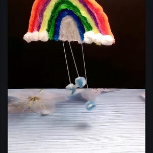 May include: A rainbow-colored yarn craft hanging from the ceiling with white cotton clouds and blue yarn raindrops hanging below.