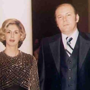 May include: A formal portrait of a man and a woman. The woman has blonde hair and is wearing a dark dress with a geometric pattern. The man is wearing a black suit with a striped tie. They are standing in front of a white pillar.