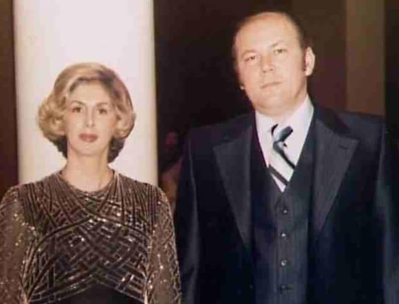 May include: A formal portrait of a man and a woman. The woman has blonde hair and is wearing a dark dress with a geometric pattern. The man is wearing a black suit with a striped tie. They are standing in front of a white pillar.