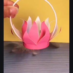 May include: A pink and white paper flower with a white paper handle. The flower is shaped like a lotus.