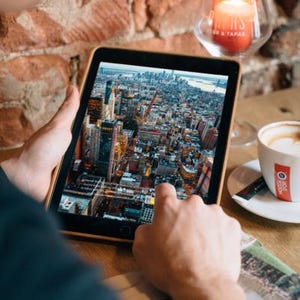 Op de afbeelding: Een tablet toont een gedetailleerd luchtfoto van een stad, waarschijnlijk New York. De tablet wordt boven een houten tafel gehouden, met een brandende kaars in een glazen houder en een kop koffie in de buurt. De bakstenen muur op de achtergrond voegt warmte toe.