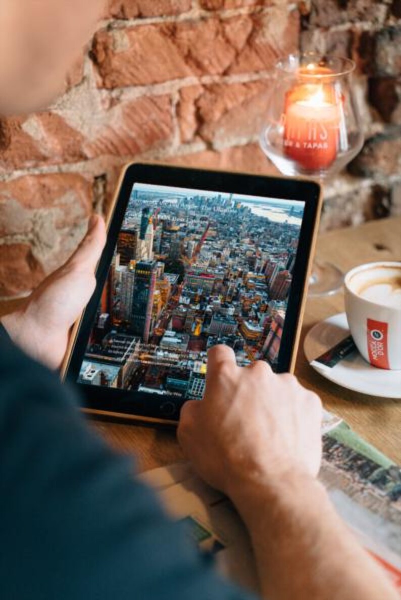 Op de afbeelding: Een tablet toont een gedetailleerd luchtfoto van een stad, waarschijnlijk New York. De tablet wordt boven een houten tafel gehouden, met een brandende kaars in een glazen houder en een kop koffie in de buurt. De bakstenen muur op de achtergrond voegt warmte toe.