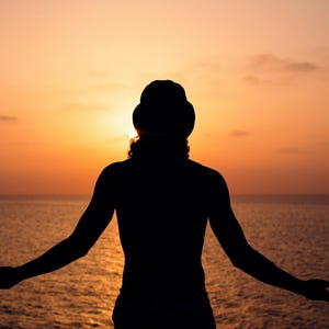 Pu&ograve; includere: Silhouette di una persona con le braccia tese, rivolta verso un tramonto sull'oceano. Il cielo &egrave; un gradiente di arancione e giallo, con la luce del sole parzialmente oscurata. L'acqua &egrave; calma e riflette i colori del cielo.