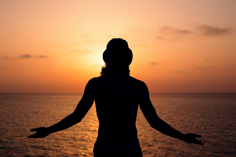 Pu&ograve; includere: Silhouette di una persona con le braccia tese, rivolta verso un tramonto sull'oceano. Il cielo &egrave; un gradiente di arancione e giallo, con la luce del sole parzialmente oscurata. L'acqua &egrave; calma e riflette i colori del cielo.