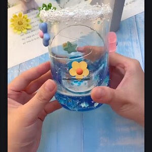 May include: A clear glass jar with a blue and white glitter design. The jar has a white lace trim around the top and is decorated with small, colorful flowers. The jar is being held by two hands.