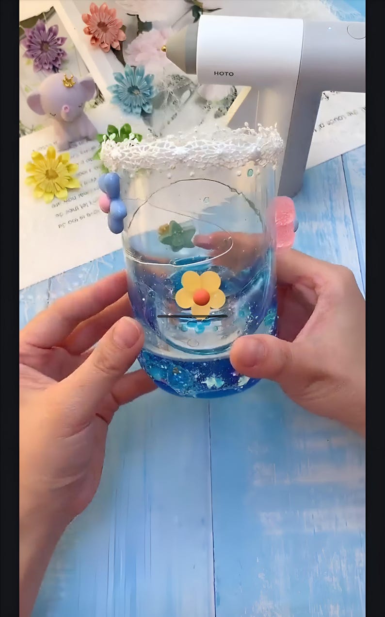 May include: A clear glass jar with a blue and white glitter design. The jar has a white lace trim around the top and is decorated with small, colorful flowers. The jar is being held by two hands.
