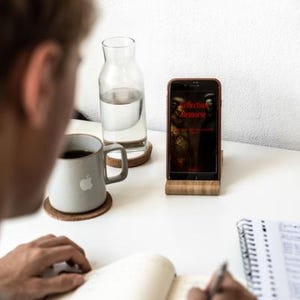 May include: A person writing in a notebook at a white desk. A smartphone in a wooden stand displays the text "Collection Remorse". A gray mug with an apple logo and a glass water bottle are also on the desk.