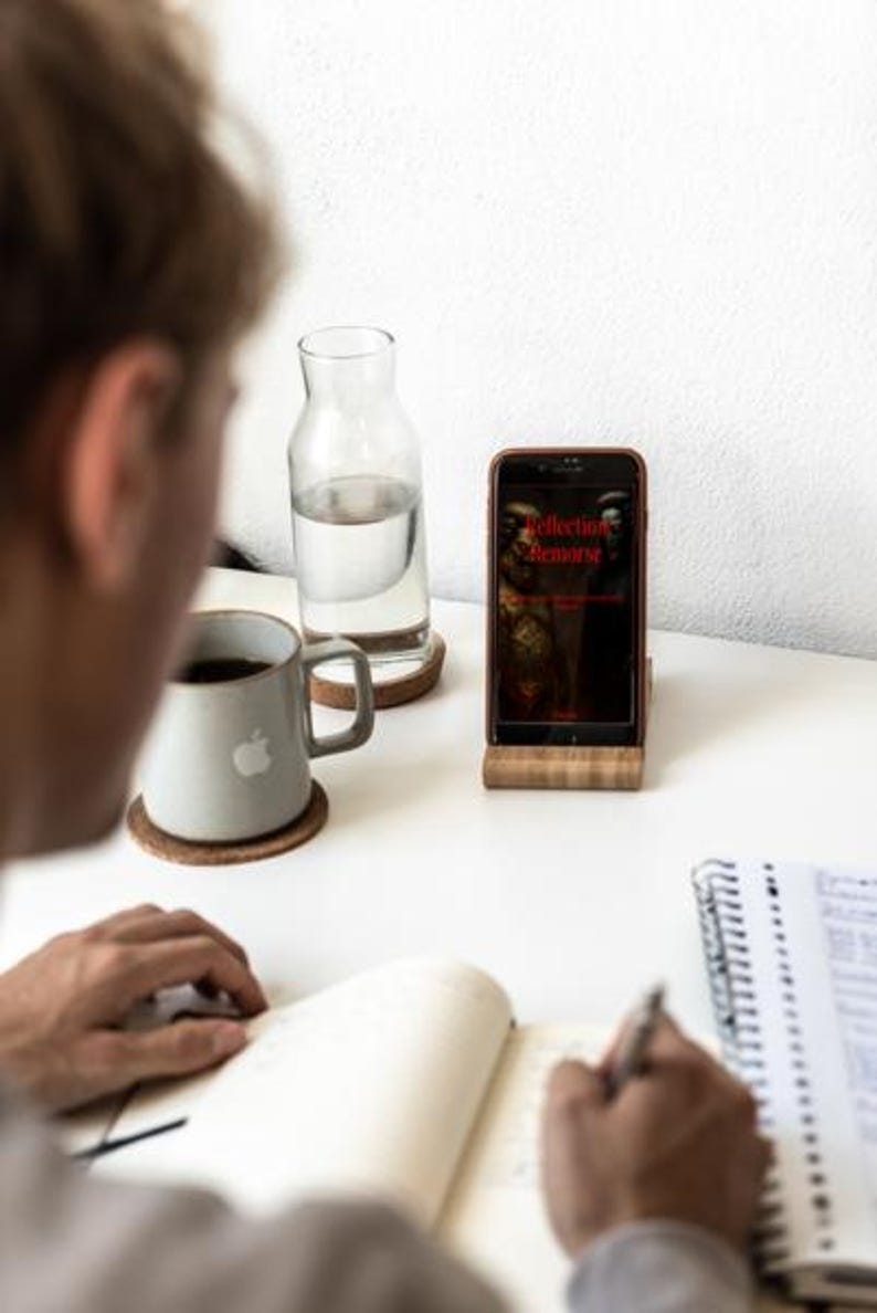 May include: A person writing in a notebook at a white desk. A smartphone in a wooden stand displays the text "Collection Remorse". A gray mug with an apple logo and a glass water bottle are also on the desk.