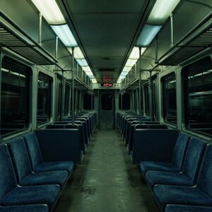 May include: The interior of a train carriage featuring rows of dark blue upholstered seats arranged on both sides of a central aisle. Overhead luggage racks run along the ceiling, which is illuminated by long, rectangular fluorescent light fixtures.