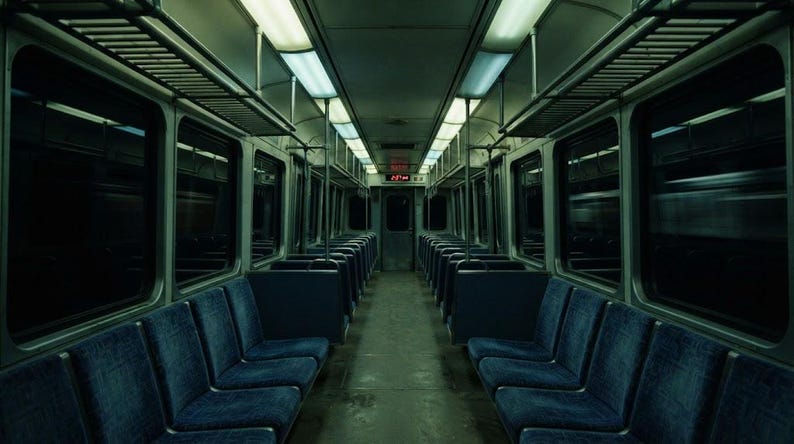 May include: The interior of a train carriage featuring rows of dark blue upholstered seats arranged on both sides of a central aisle. Overhead luggage racks run along the ceiling, which is illuminated by long, rectangular fluorescent light fixtures.