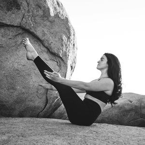 Pu&ograve; includere: Una persona in abbigliamento sportivo nero esegue una posa yoga all'aperto. La persona &egrave; seduta su una roccia, con le gambe estese verso l'alto e le braccia tese. Lo sfondo presenta grandi formazioni rocciose.